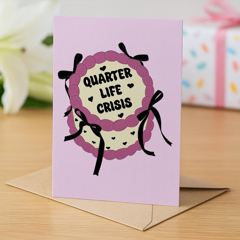May include: A greeting card with a pink background and a cake design. The cake has a cream-colored center with the words "Quarter Life Crisis" and small black hearts. The cake is decorated with purple frosting and black bows.