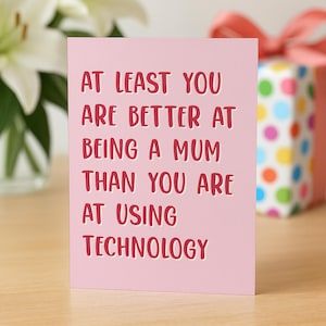 Puede incluir: Una tarjeta rosa con el texto "AT LEAST YOU ARE BETTER AT BEING A MUM THAN YOU ARE AT USING TECHNOLOGY" en rojo. Una caja de regalo con una cinta rosa está en el fondo.
