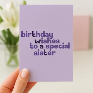 May include: A lavender birthday card with the text "birthday wishes to a special sister" in shades of purple. The card is held by a hand with a white painted fingernail. A vase of white flowers is in the background.