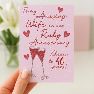 Può includere: Un biglietto d'auguri rettangolare con sfondo rosa chiaro, cuori rossi e due flûte da champagne. Il testo recita: To my Amazing Wife on our Ruby Anniversary Cheers to 40 years! Il design include piccoli cuori bianchi come decorazione.