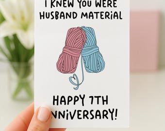 May include: A white greeting card featuring a pink yarn ball and a blue yarn ball leaning against each other with a heart-shaped yarn strand. The text reads: I KNEW YOU WERE HUSBAND MATERIAL HAPPY 7TH ANNIVERSARY! in black, rounded, handwritten-style font.