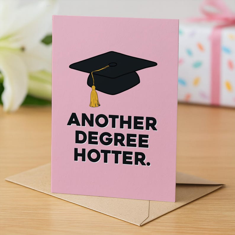 Pu&ograve; includere: Un biglietto d'auguri rosa con un tocco di laurea nero e una nappa gialla. Il testo sul biglietto recita "ANOTHER DEGREE HOTTER". Il biglietto &egrave; posizionato in verticale su una busta marrone chiaro, con un design pulito e semplice.