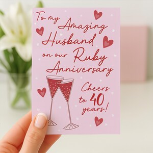 Könnte beinhalten: Eine hellrosa Grußkarte mit dunkelrotem Text, der To my Amazing Husband on our Ruby Anniversary und Cheers to 40 years! lautet. Das Design zeigt zwei Champagnergläser mit roter Flüssigkeit und mehrere kleine rote Herzen.