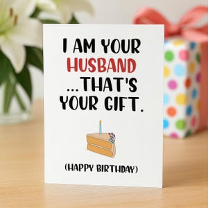 May include: A white greeting card with black and red text that reads I AM YOUR HUSBAND ... THAT'S YOUR GIFT. (HAPPY BIRTHDAY). Below the text is a simple illustration of a slice of cake with a single blue candle and colorful sprinkles.
