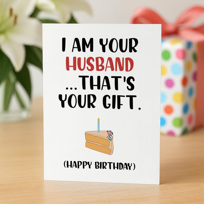 May include: A white greeting card with black and red text that reads I AM YOUR HUSBAND ... THAT'S YOUR GIFT. (HAPPY BIRTHDAY). Below the text is a simple illustration of a slice of cake with a single blue candle and colorful sprinkles.