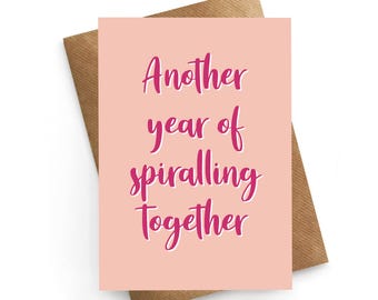May include: A greeting card with the text "Another year of spiralling together" in pink script on a light pink background. The card is on a brown envelope.