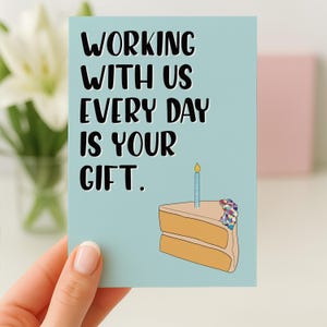 Pode incluir: Cartão azul claro com o texto "WORKING WITH US EVERY DAY IS YOUR GIFT." Uma fatia de bolo com uma vela acesa é ilustrada no canto inferior direito. O cartão é segurado por uma mão.