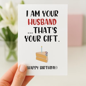 May include: A white greeting card with black and red text that reads I AM YOUR HUSBAND... THAT'S YOUR GIFT. Below the text is a drawing of a slice of cake with a single blue candle, followed by the text (HAPPY BIRTHDAY) in black.
