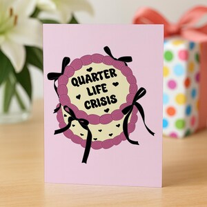 May include: A pink greeting card features a cake design with the text "Quarter Life Crisis." The cake has a light yellow center, a pink frosting border, and black bow accents. Small black heart shapes are scattered around the cake.