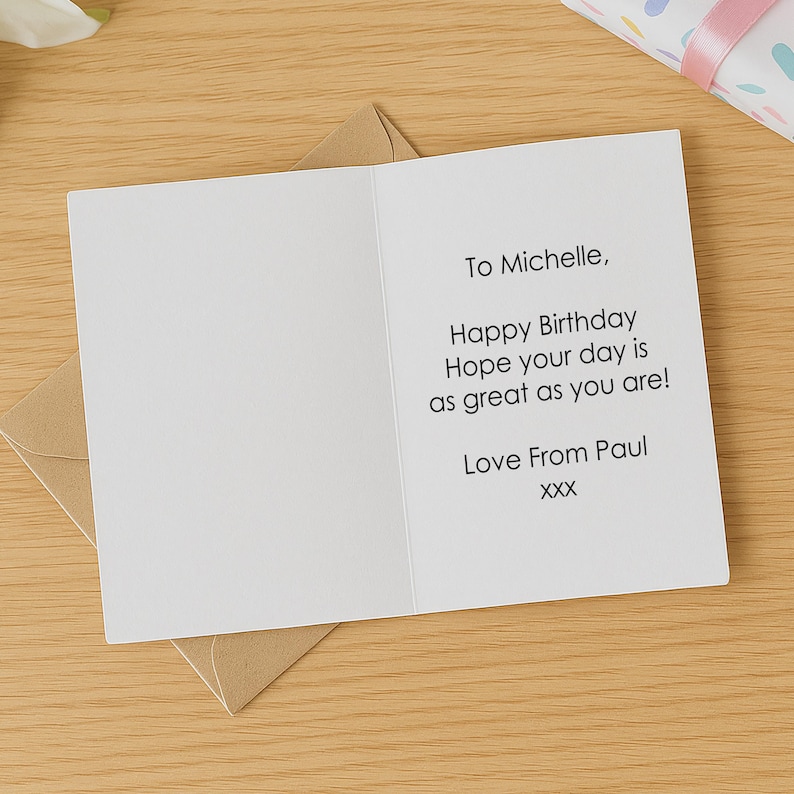 May include: A white birthday card with the message "To Michelle, Happy Birthday Hope your day is as great as you are! Love From Paul XXX" is open on a wooden surface, resting on a brown envelope.