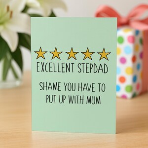May include: A light green greeting card with the text "EXCELLENT STEPDAD" and "SHAME YOU HAVE TO PUT UP WITH MUM" in black. The card has five gold star outlines above the text. A gift box with colorful polka dots is in the background.