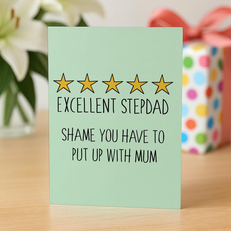 May include: A light green greeting card with the text "EXCELLENT STEPDAD" and "SHAME YOU HAVE TO PUT UP WITH MUM" in black. The card has five gold star outlines above the text. A gift box with colorful polka dots is in the background.
