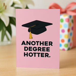 Pu&ograve; includere: Un biglietto d'auguri rettangolare con sfondo rosa chiaro raffigurante un tocco di laurea nero con una nappa gialla. Sotto il tocco, il testo in grassetto nero a caratteri maiuscoli recita: ANOTHER DEGREE HOTTER. Il design &egrave; pulito e piatto.