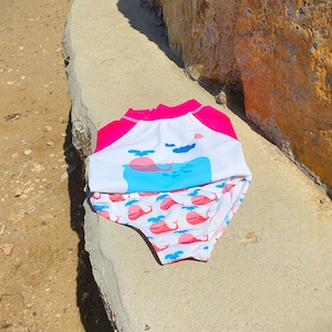 May include: Pink and white swimsuit with a whale print. The top has a pink and white color block design. The bottom has a white background with blue whales and pink accents.