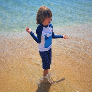 May include: A young child wearing a white and blue rash guard with a whale graphic and blue shorts is standing in shallow water on a sandy beach.