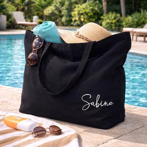 May include: Black tote bag with the name "Sabine" in white script. The bag contains a rolled turquoise towel, a straw hat, and sunglasses. A beach towel, sunglasses, and sunscreen are in front of the bag.