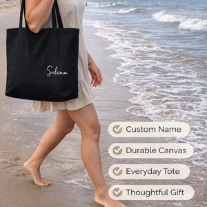 May include: A black tote bag with the name "Selena" in white script is carried by a person walking on a beach. The bag has black handles and the words "Personalized Beach Tote Bag" are in the background.