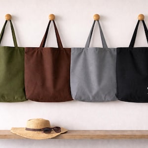 May include: Four canvas tote bags in olive green, brown, grey, and black, suspended from wooden pegs. The black bag features the name "Sabine" in white script. A straw hat and sunglasses sit on a wooden shelf.