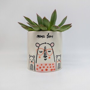 May include: White ceramic planter with a hand-drawn illustration of a mama bear wearing glasses and two baby bears. The planter has pink polka dots and the text "mama bear" is written on the front. The planter is filled with a green succulent plant.