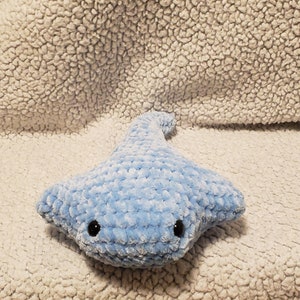 May include: A light blue crocheted stingray plush toy with black eyes. The toy is sitting on a white fluffy surface.