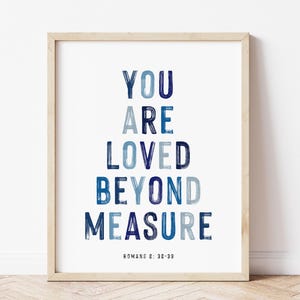 Op de afbeelding: Een ingelijste kunstprint met de tekst YOU ARE LOVED BEYOND MEASURE in een gestapeld lettertype met penseelstreek-effect in verschillende blauwtinten. Onder de hoofdtekst staat in kleinere zwarte letters ROMANS 8: 38-39. De lijst is van licht hout.