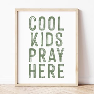Op de afbeelding: Een ingelijste print met de woorden "COOL KIDS PRAY HERE" in groene penseelstreken. De print zit in een licht houten lijst en wordt weergegeven tegen een witte muur. De vloer is licht van kleur.