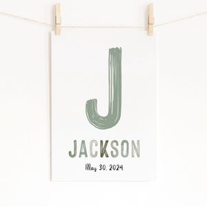 Op de afbeelding: Een witte print met de naam "Jackson" in groene en grijze letters, met de datum "May 30, 2024" eronder. Een grote groene letter "J" staat bovenaan. De print hangt aan een touwtje met houten wasknijpers.