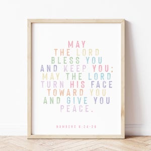 Op de afbeelding: Een ingelijste print met een licht houten frame. De print bevat de tekst "MAY THE LORD BLESS YOU AND KEEP YOU; MAY THE LORD TURN HIS FACE TOWARD YOU AND GIVE YOU PEACE." in pastelkleuren. De tekst "NUMBERS 6:24-26" staat onderaan.