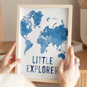 Op de afbeelding: Een ingelijste print met een blauwe aquarel wereldkaart. De woorden "LITTLE EXPLORER" staan in het blauw onder de kaart. De lijst is licht van kleur.