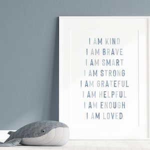 以下が含まれることがあります： 「I AM KIND, BRAVE, SMART, STRONG, GRATEFUL, HELPFUL, ENOUGH, LOVED」という言葉が青い文字で書かれたフレーム入りのプリント。灰色のクジラのぬいぐるみのおもちゃが、青い壁を背景に、プリントの前に床に置かれています。