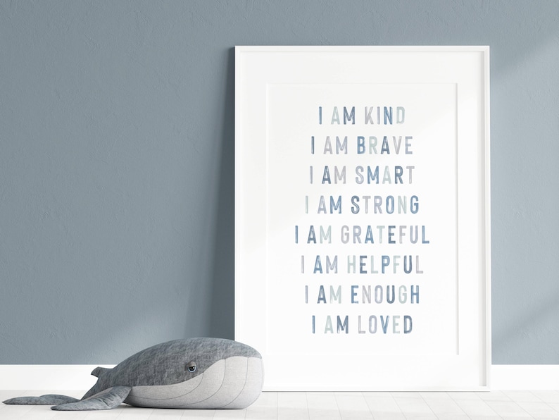 以下が含まれることがあります： 「I AM KIND, BRAVE, SMART, STRONG, GRATEFUL, HELPFUL, ENOUGH, LOVED」という言葉が青い文字で書かれたフレーム入りのプリント。灰色のクジラのぬいぐるみのおもちゃが、青い壁を背景に、プリントの前に床に置かれています。