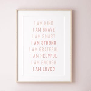 May include: A framed print with the words "I AM KIND, I AM BRAVE, I AM SMART, I AM STRONG, I AM GRATEFUL, I AM HELPFUL, I AM ENOUGH, I AM LOVED" in a soft pink font. The print is in a white mat and a light wood frame.