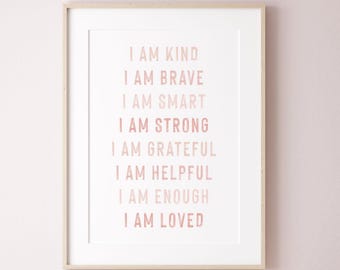 May include: A framed print with the words "I AM KIND, I AM BRAVE, I AM SMART, I AM STRONG, I AM GRATEFUL, I AM HELPFUL, I AM ENOUGH, I AM LOVED" in a soft pink font. The print is in a white mat and a light wood frame.