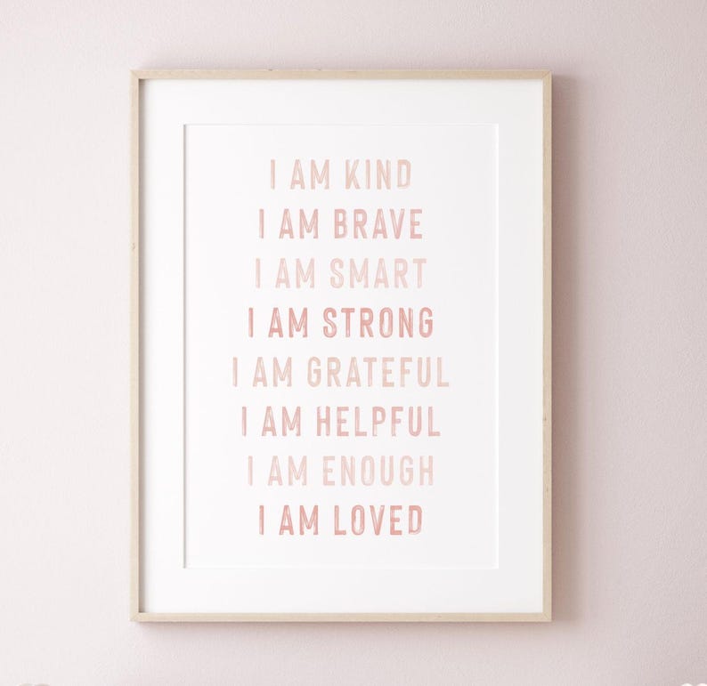 May include: A framed print with the words "I AM KIND, I AM BRAVE, I AM SMART, I AM STRONG, I AM GRATEFUL, I AM HELPFUL, I AM ENOUGH, I AM LOVED" in a soft pink font. The print is in a white mat and a light wood frame.