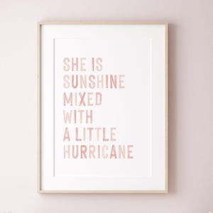Pode incluir: Impressão emoldurada com a citação "SHE IS SUNSHINE MIXED WITH A LITTLE HURRICANE" em letras rosa. O texto está centrado em um fundo branco, dentro de uma moldura de madeira clara. Adequado para um quarto infantil.