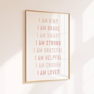 May include: A framed print with a gold-colored frame hangs on a light pink wall. The print features the phrases "I AM KIND, BRAVE, SMART, STRONG, GRATEFUL, HELPFUL, ENOUGH, LOVED" in a soft pink font. The wall is lit by natural light.
