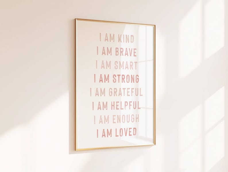 May include: A framed print with a gold-colored frame hangs on a light pink wall. The print features the phrases "I AM KIND, BRAVE, SMART, STRONG, GRATEFUL, HELPFUL, ENOUGH, LOVED" in a soft pink font. The wall is lit by natural light.