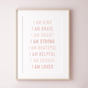 Op de afbeelding: Een ingelijste print met de woorden "I AM KIND, I AM BRAVE, I AM SMART, I AM STRONG, I AM GRATEFUL, I AM HELPFUL, I AM ENOUGH, I AM LOVED" in roze. De print zit in een witte passe-partout en een licht houten frame.