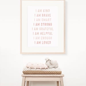 May include: A framed print with the words "I AM KIND, I AM BRAVE, I AM SMART, I AM STRONG, I AM GRATEFUL, I AM HELPFUL, I AM ENOUGH, I AM LOVED" in pink. Below the frame, a stack of blankets and a small stuffed bunny sit on a wooden stool.