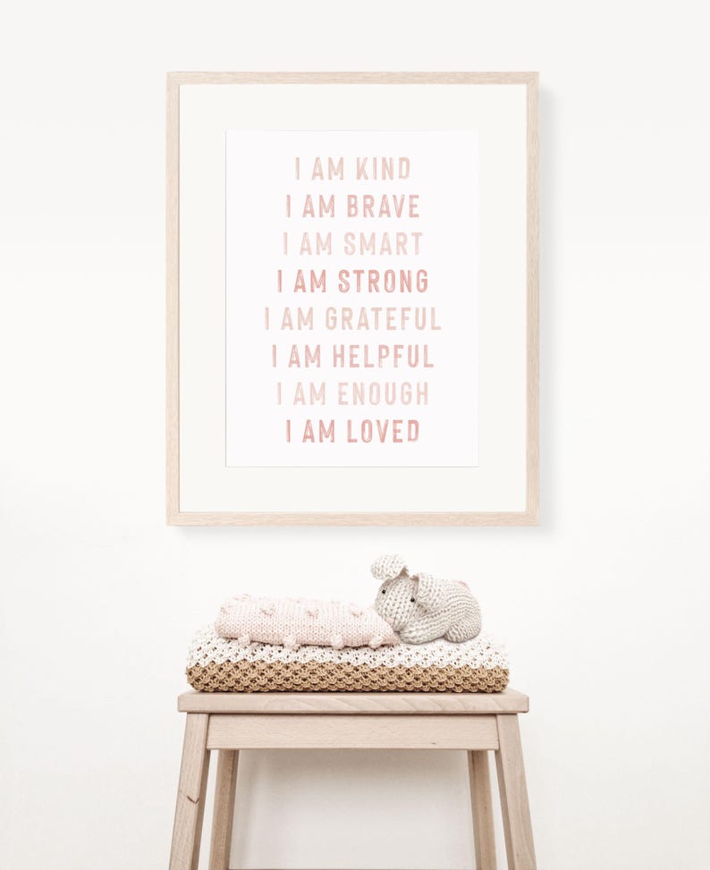 May include: A framed print with the words "I AM KIND, I AM BRAVE, I AM SMART, I AM STRONG, I AM GRATEFUL, I AM HELPFUL, I AM ENOUGH, I AM LOVED" in pink. Below the frame, a stack of blankets and a small stuffed bunny sit on a wooden stool.