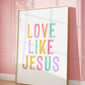 Op de afbeelding: Een ingelijste print met de tekst "LOVE LIKE JESUS" in kleurrijke letters. De tekst gebruikt tinten geel, oranje, roze, paars en turquoise. Het kunstwerk wordt gepresenteerd tegen een roze muur en een houten vloer.
