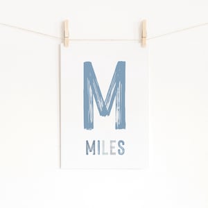 Op de afbeelding: Een witte print met een grote blauwe letter "M" en de naam "MILES" in een lichtere blauwe letter. De print wordt met houten wasknijpers aan een touwtje tegen een witte achtergrond gehangen.
