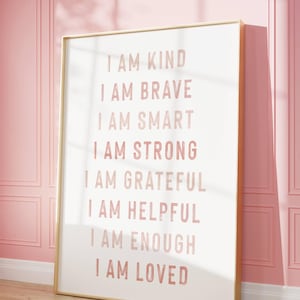 May include: A framed print with a gold border displays the words "I AM KIND, I AM BRAVE, I AM SMART, I AM STRONG, I AM GRATEFUL, I AM HELPFUL, I AM ENOUGH, I AM LOVED" in pink lettering on a white background. The print leans against a pink wall.