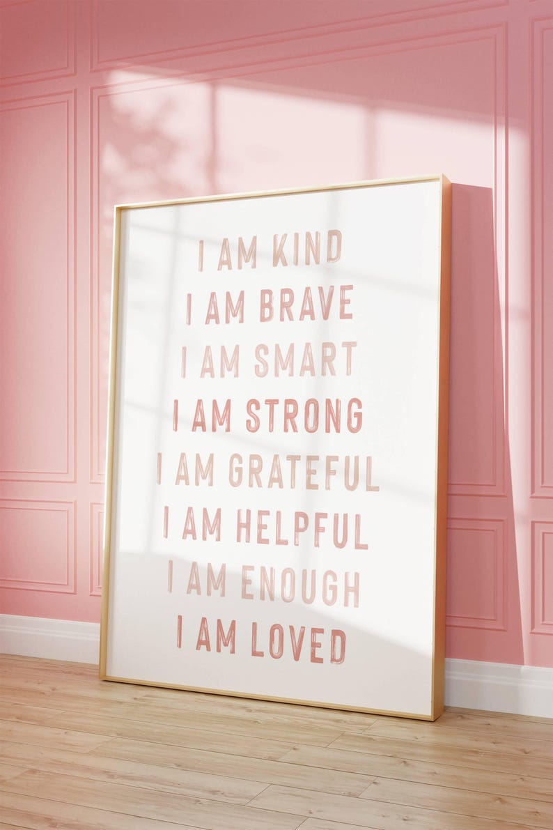 May include: A framed print with a gold border displays the words "I AM KIND, I AM BRAVE, I AM SMART, I AM STRONG, I AM GRATEFUL, I AM HELPFUL, I AM ENOUGH, I AM LOVED" in pink lettering on a white background. The print leans against a pink wall.