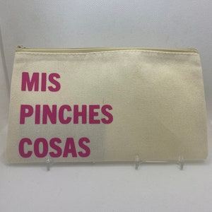 May include: A beige canvas zippered pouch with the words "MIS PINCHES COSAS" printed in bright pink. The pouch is rectangular and appears to be empty. The background is a plain white surface.