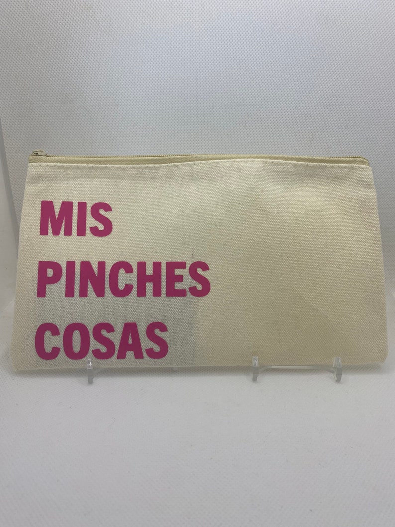 May include: A beige canvas zippered pouch with the words "MIS PINCHES COSAS" printed in bright pink. The pouch is rectangular and appears to be empty. The background is a plain white surface.