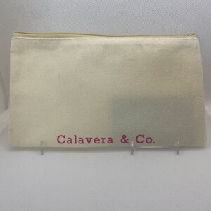 May include: A cream-colored canvas zippered pouch with the text "Calavera & Co." in pink. The pouch is rectangular and appears to be empty. The zipper is a light beige color.