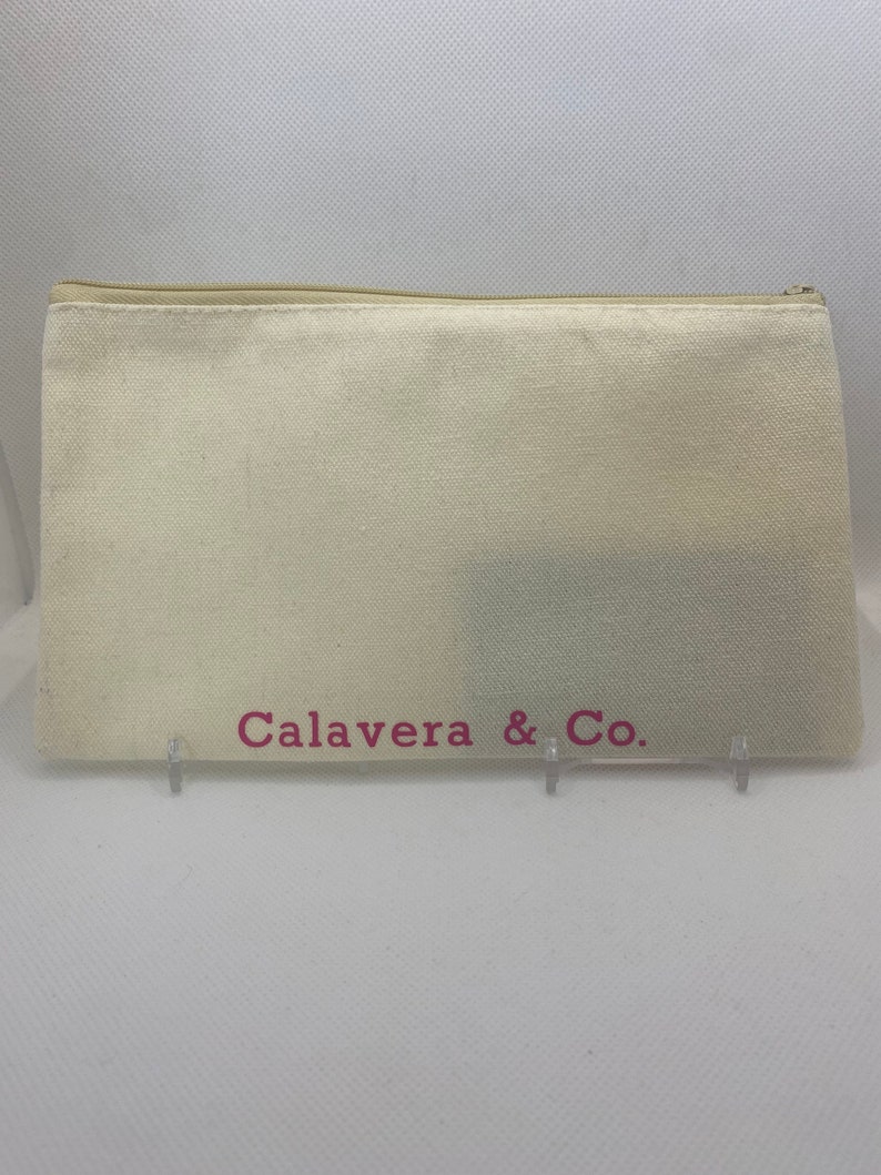 May include: A cream-colored canvas zippered pouch with the text "Calavera & Co." in pink. The pouch is rectangular and appears to be empty. The zipper is a light beige color.