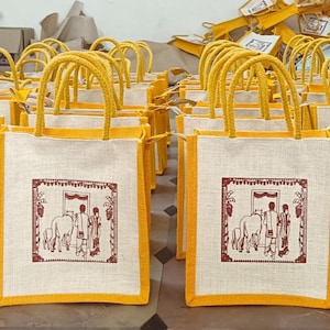 Può includere: Un gruppo di piccole borse di juta gialle e bianche con manici. Ogni borsa ha un disegno rosso e bianco di un cavallo e persone. Le borse sono allineate in una fila.
