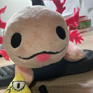 Könnte beinhalten: Ein Plüschtier-Axolotl mit rosa Kiemen und einer rosa Zunge. Daneben befindet sich ein gelbes dreieckiges Plüschtier mit schwarzem Auge und Fliege sowie ein kleines schwarzes Plüschtier.